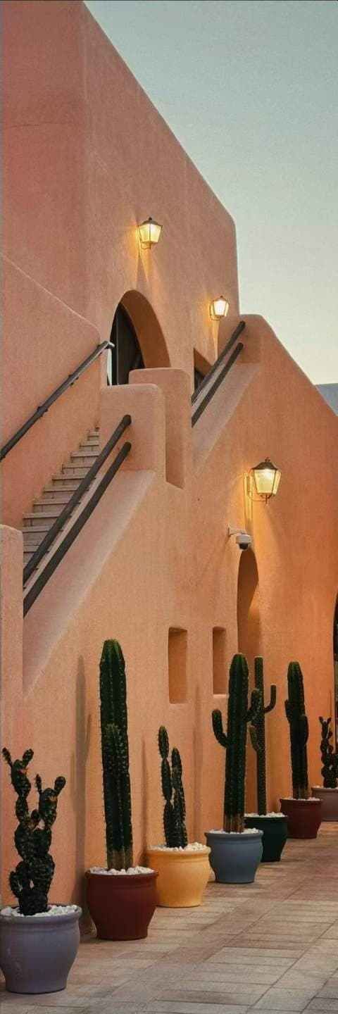 An external stair on a terracotta building at dusk, lanterns lit, potted cacti along the base.